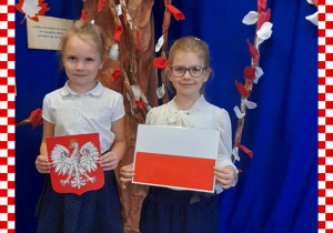 Jula i Klara w odświętnym ubraniu trzymają w rączkach godło i flagę Polski, w tle dekoracja okolicznościowa